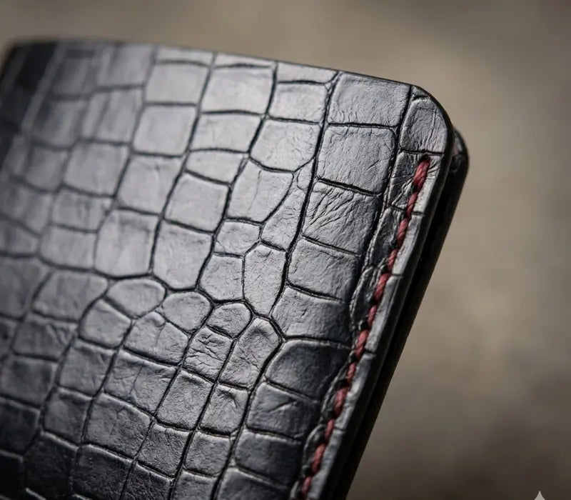 Close-up of a black leather wallet with crocodile texture and red stitching on a blurred background.