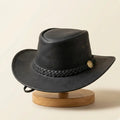 Black handmade full-grain leather cowboy hat with braided band and brass hardware on a beige background