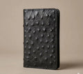 Black genuine ostrich leather handmade long wallet with brass snap closures on a beige background