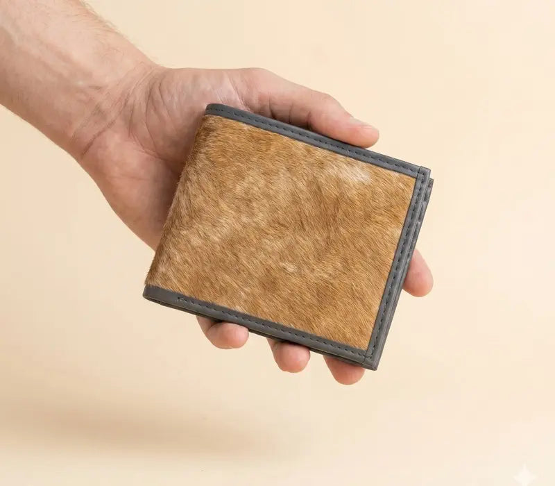 Tan hair-on cowhide handmade leather bifold wallet with grey trim held in a hand